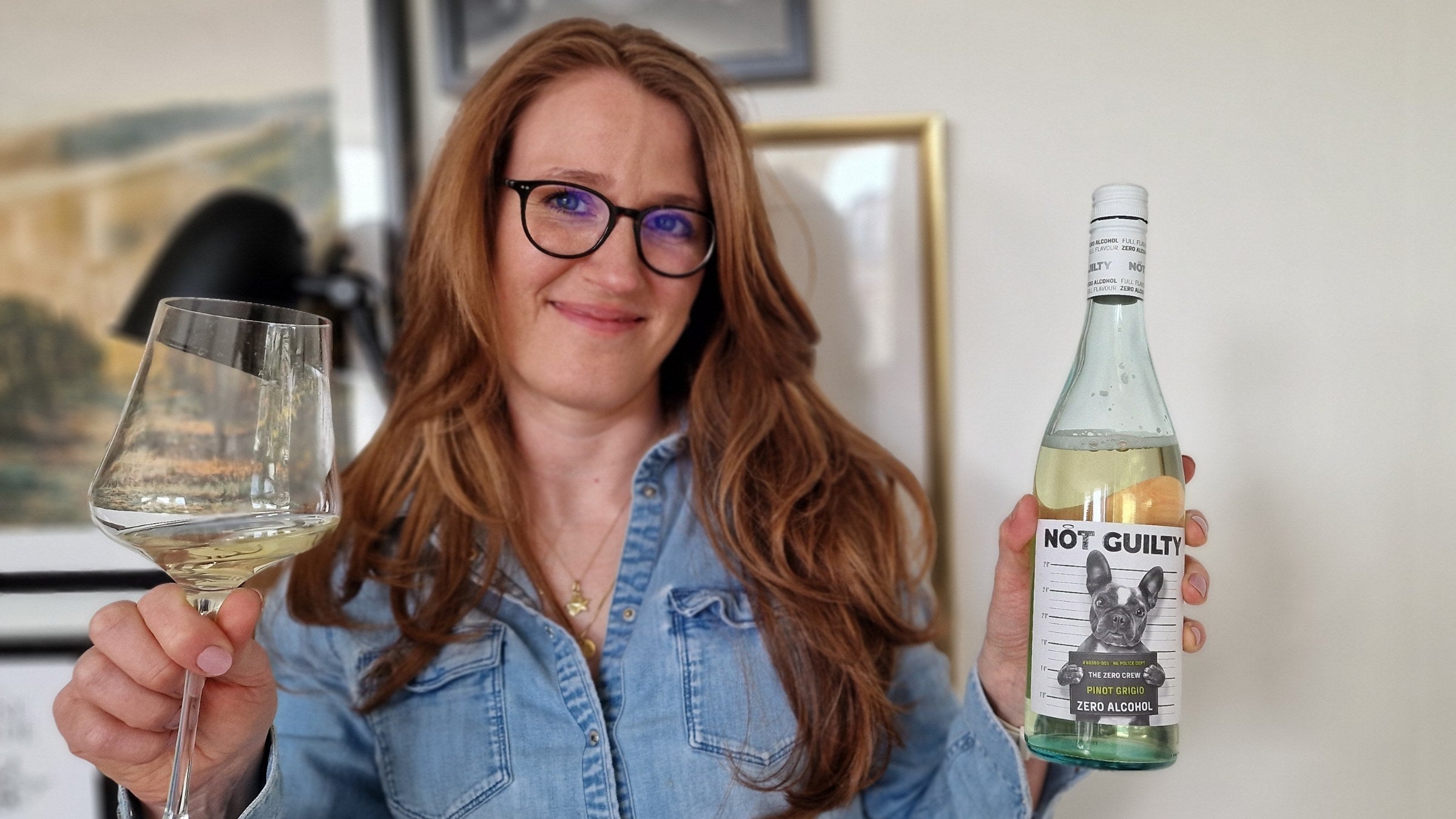 Margot van Lieshout holding a bottle of NOT GUILTY zero-alcohol Pinot Grigio and a glass of white wine, smiling to camera in a denim shirt.