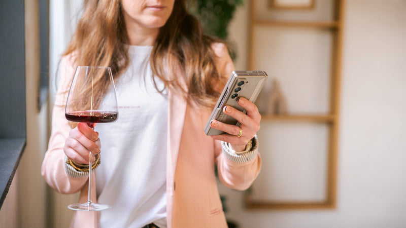 A person relaxed at home with a phone in one hand and a glass of wine nearby, searching in warm natural light