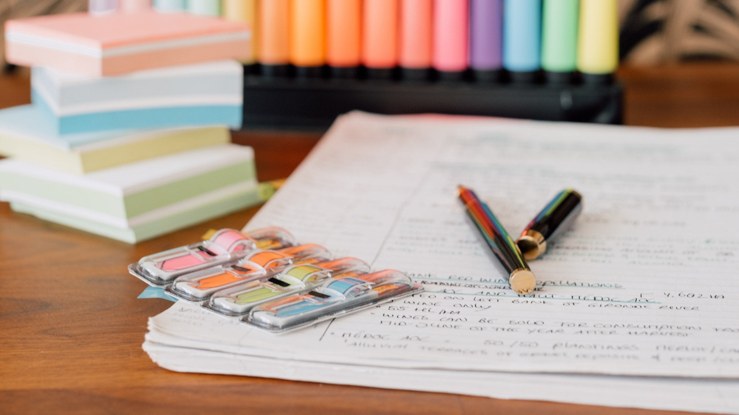 An open WSET study notebook filled with handwriting on a warm wooden desk, beside a watercolour paint palette used as a colour key, a stack of pastel sticky notes and two pens, in soft natural light.