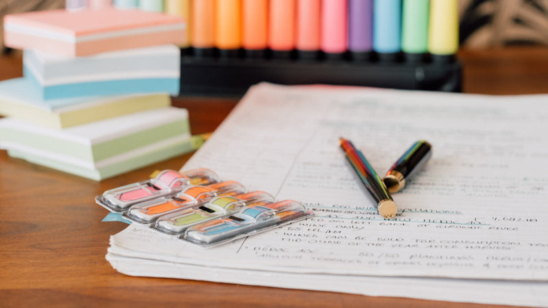 An open WSET study notebook filled with handwriting on a warm wooden desk, beside a watercolour paint palette used as a colour key, a stack of pastel sticky notes and two pens, in soft natural light.