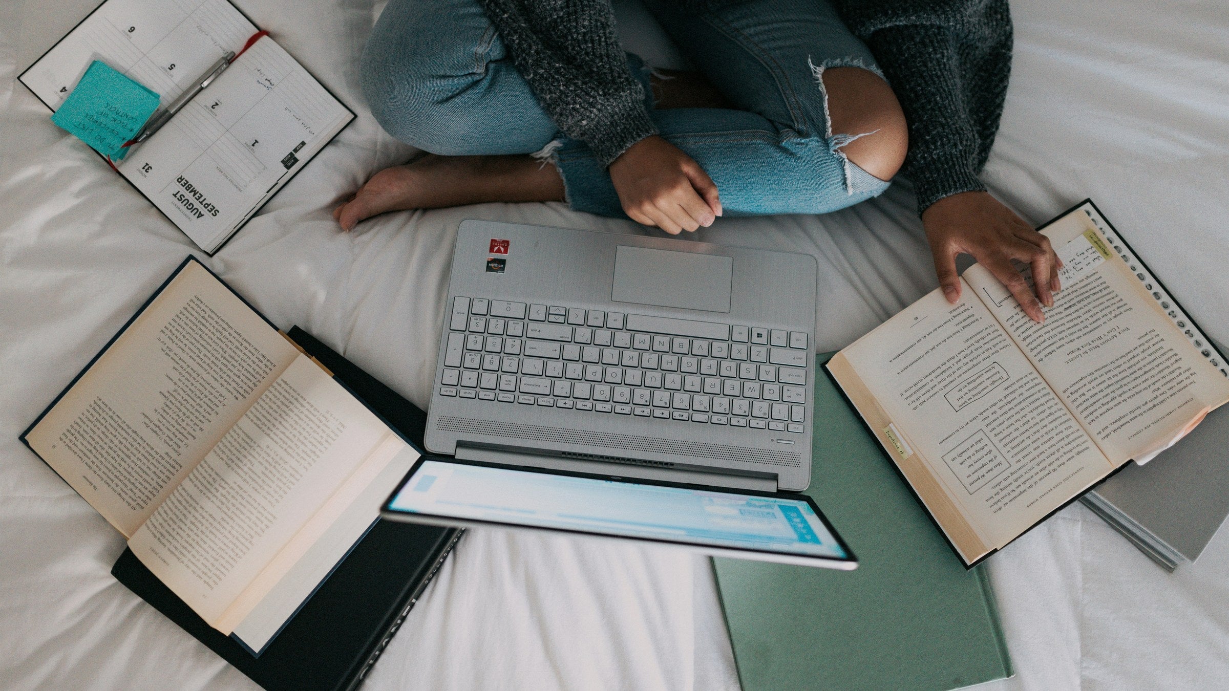 Overhead view of a study setup on a white bed, an open laptop surrounded by open books, a tablet, a green notebook and a spiral planner with sticky notes.