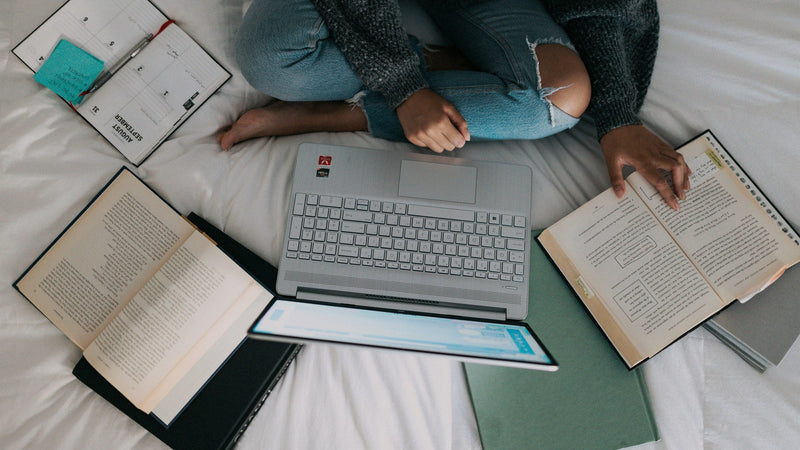 Overhead view of a study setup on a white bed, an open laptop surrounded by open books, a tablet, a green notebook and a spiral planner with sticky notes.