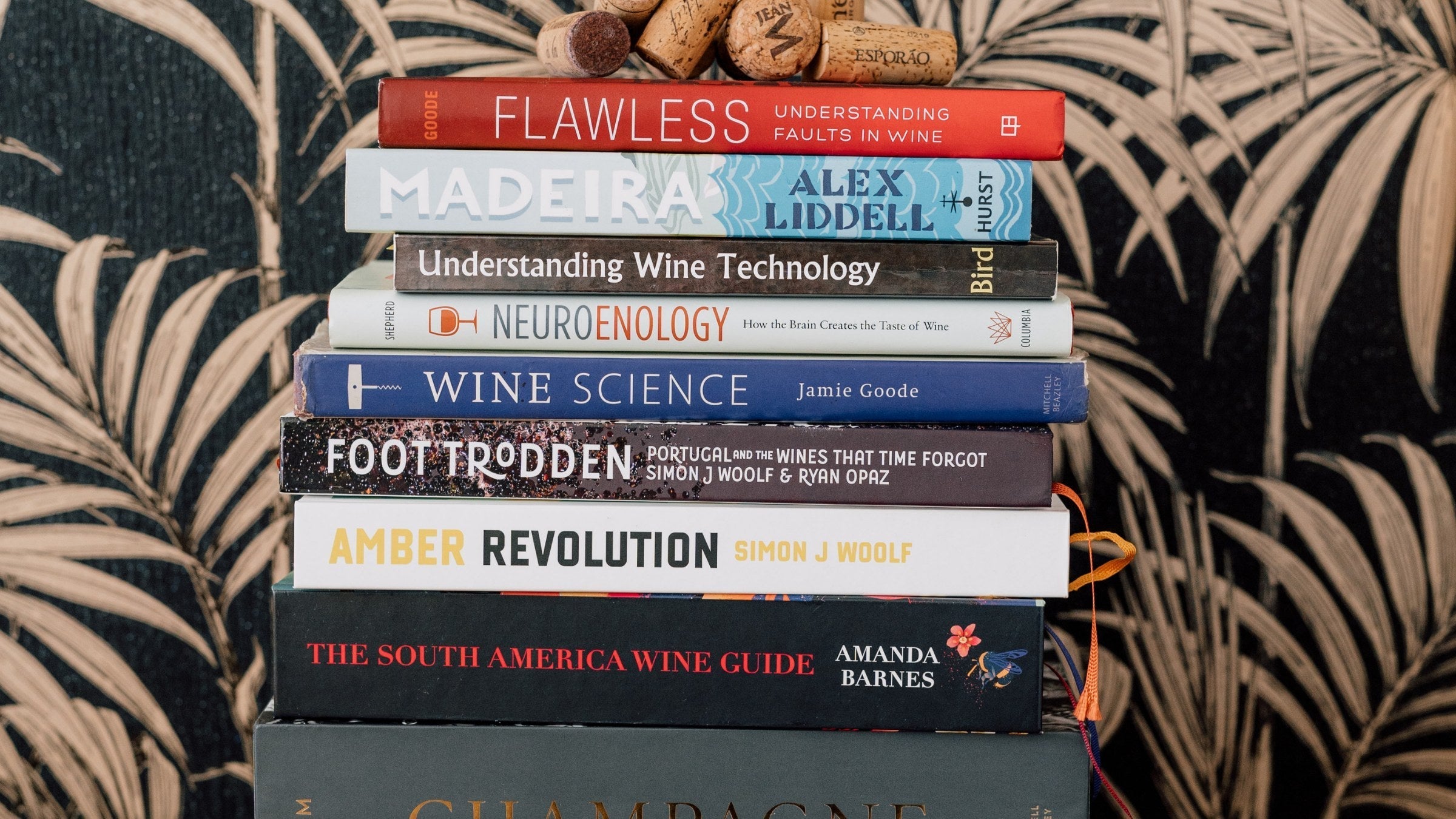 A neat stack of wine study books, including Wine Science, Amber Revolution, Understanding Wine Technology and The South America Wine Guide, topped with two corks, against dark palm-patterned wallpaper.