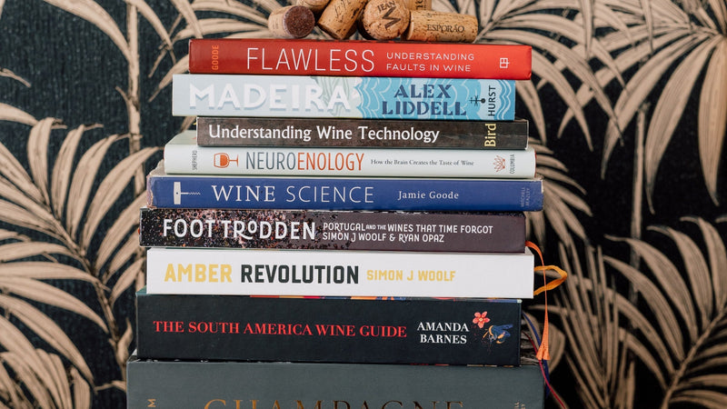 A neat stack of wine study books, including Wine Science, Amber Revolution, Understanding Wine Technology and The South America Wine Guide, topped with two corks, against dark palm-patterned wallpaper.