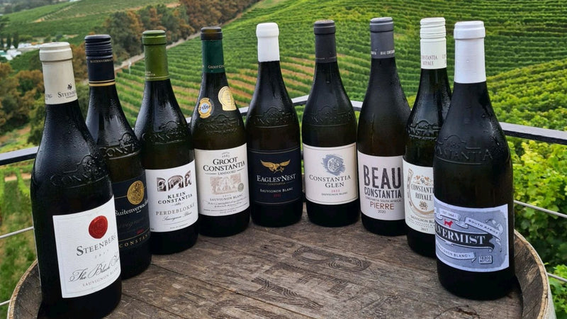 Nine bottles of Constantia Sauvignon Blanc lined up on a barrel above the vineyards, from Steenberg and Klein Constantia to Groot Constantia, Eagles' Nest, Constantia Glen, Beau Constantia and Silvermist