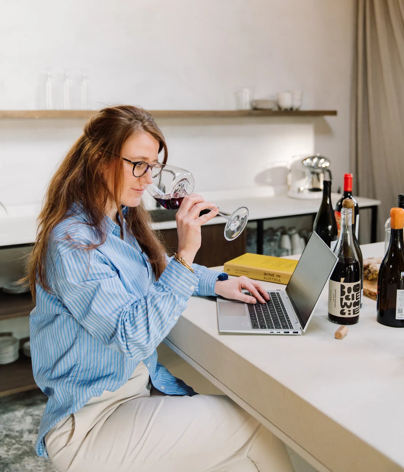 Margot van Lieshout tasting a glass of red wine while working on her laptop, wine bottles lined up on the counter